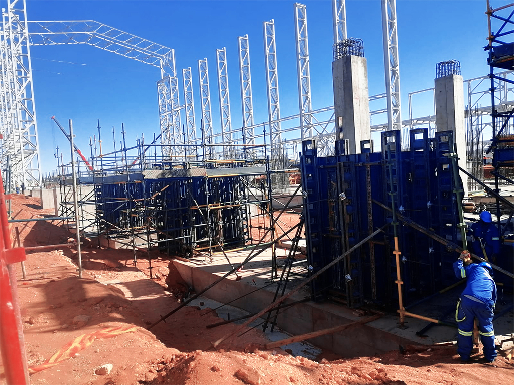 A construction site featuring multiple structural components under development. Visible are steel frameworks and scaffolding, along with concrete columns reinforced by steel rebar. Workers in blue uniforms and safety gear are actively engaged in various tasks. The ground consists of red soil, trenches, and foundational structures. The clear blue sky suggests favorable weather conditions for the ongoing work. The scene highlights the complexity and scale of the project, showcasing the various stages of building a large structure.